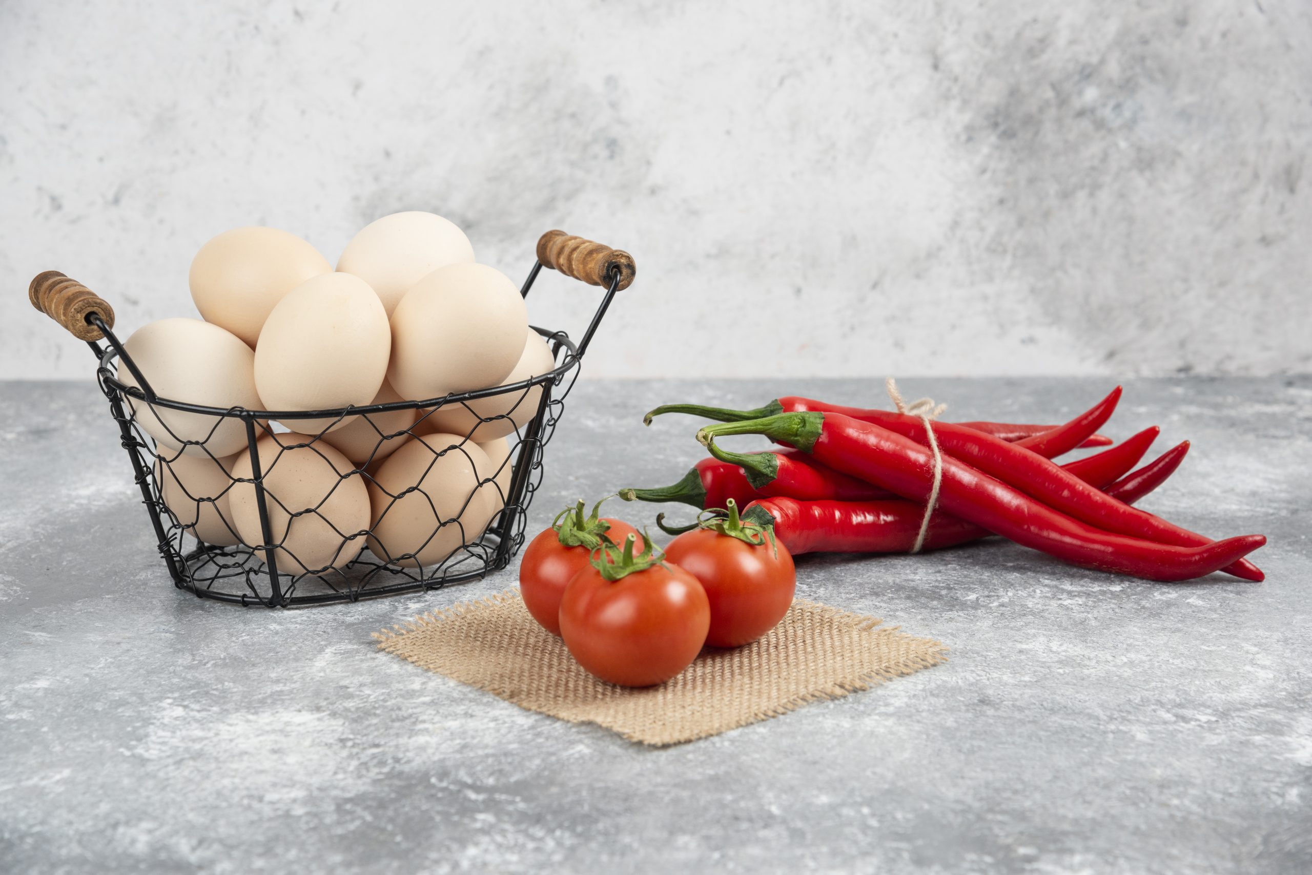 Basket of fresh uncooked eggs, chili peppers and tomatoes on marble background
